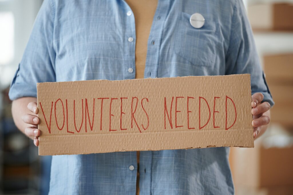 A volunteer holding a cardboard sign that reads Volunteers Needed